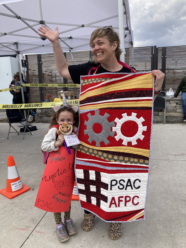 A person holding a PSAC quilt waves as her child stands next to her.