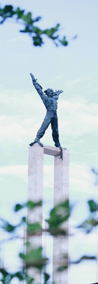 A gray and black statue of a human with arms outstretched during the daytime, pictured through trees.