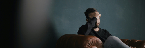 A bearded white man sits in a comfy chair, holding his head in his hand