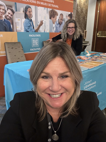 Catherine&rsquo;s smiling face is close to the camera, while Roxanne poses behind her at the JLP kiosk.
