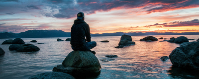 A person sits cross-legged on a rock, surrounded by water. The person is silhouetted against the sunset.
