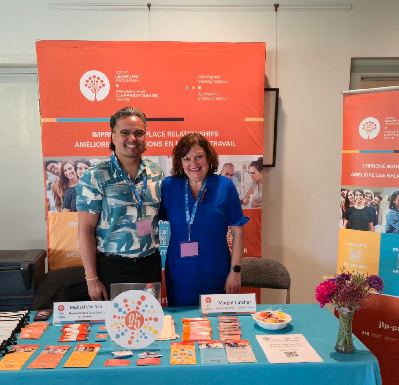 Michael et Margot sont au kiosque du PAM et sourient &agrave; la cam&eacute;ra. Sur la table devant eux se trouvent des articles promotionnels du Programme.
