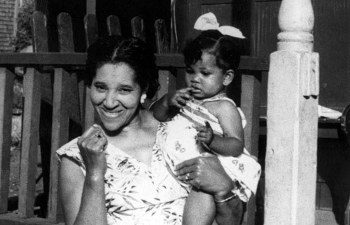 Une jeune femme noire sourit &agrave; la cam&eacute;ra. Elle se tient devant une maison et porte dans ses bras une petite fille noire. La photo, prise &agrave; Africville, est en noir et blanc.