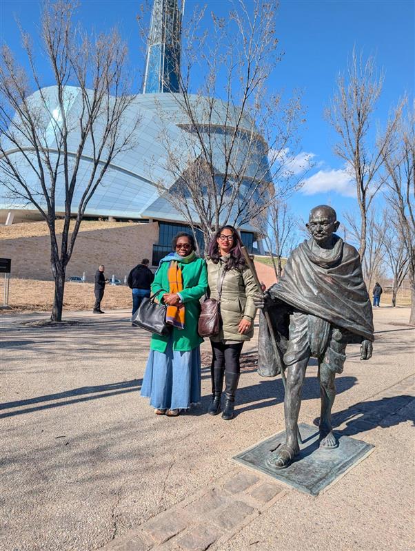 Micha&euml;lle and Anu stand next to a statue of Mahatma Gandhi, while the Human Rights Museum rises in the background. 