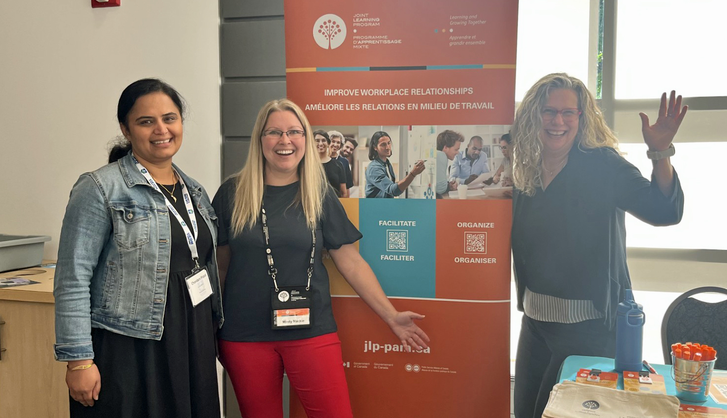 Chandani, Mindy et Roxanne sourient et font signe de la main devant la banni&egrave;re du PAM.